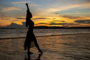Silhouette of a woman raising arms on a beach during a dramatic sunset.